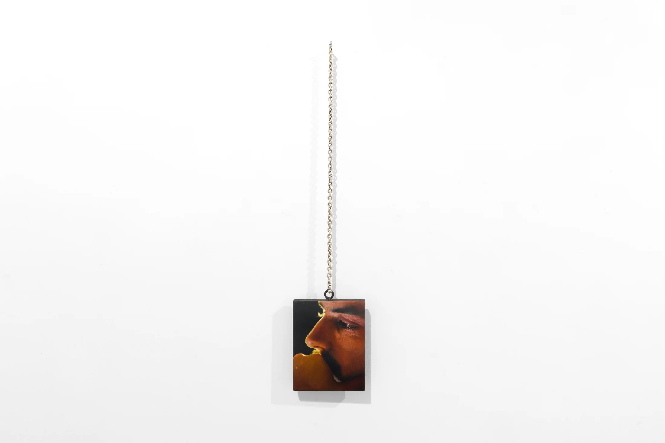 a painting by Ki Yoong of a close up of a male face against a dark background which hangs from a silver necklace