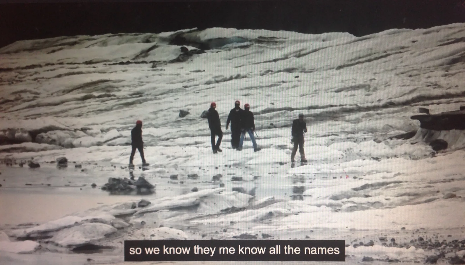 An image of Cath Campbell's film 'for quartets' - 5 figures wearing black walk across an icy landscape - the words "so we know they me know all the names" are written across the bottom as a subtitle.