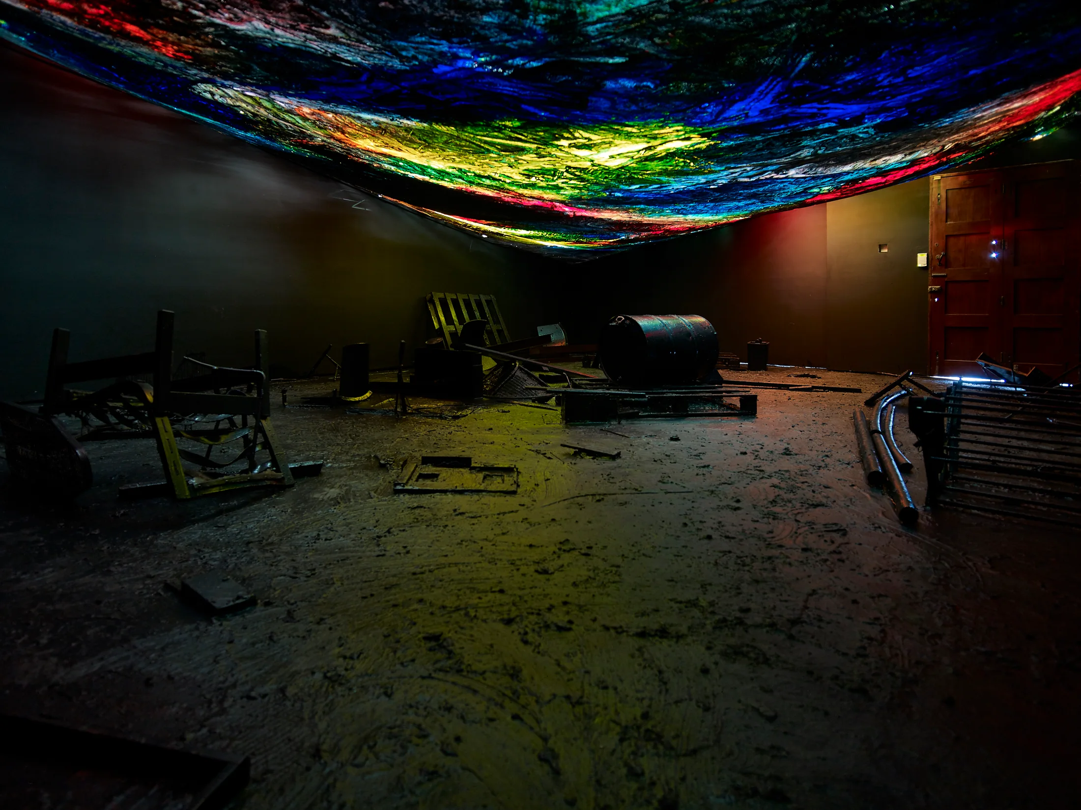 A installation by Hugo Canoilas consisting of darkened room with objects covered in black latex, and a low painted colourful canvas as ceiling