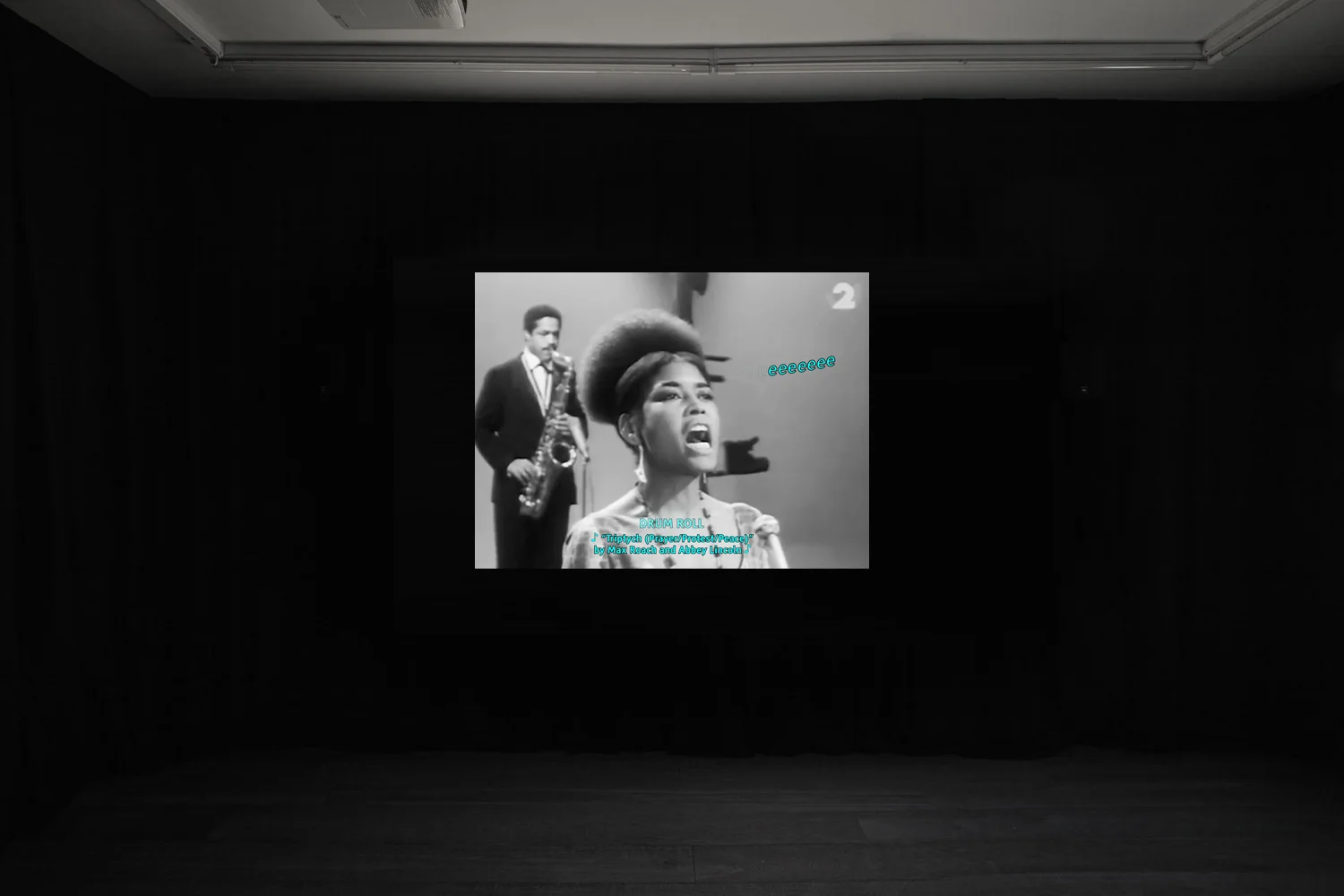 An install shot of Rosa-Johan Uddoh's film at Workplace in London. A black and white image of a saxophonist and singer performing. Over the top is blue text 'eeeeeee' describing the sound.