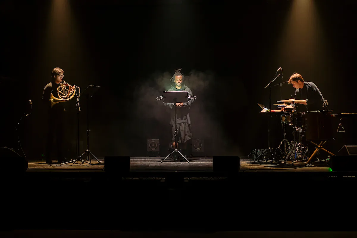 Simeon Barclay performing with two musicians on a dark stage