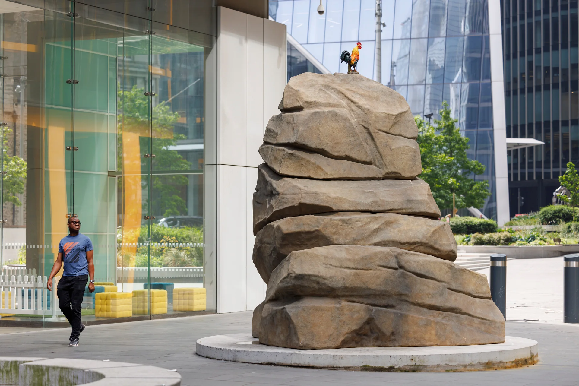 A large sculpture of a boulder with a cockerel on the top situated in a square surrounded by glass buildings 