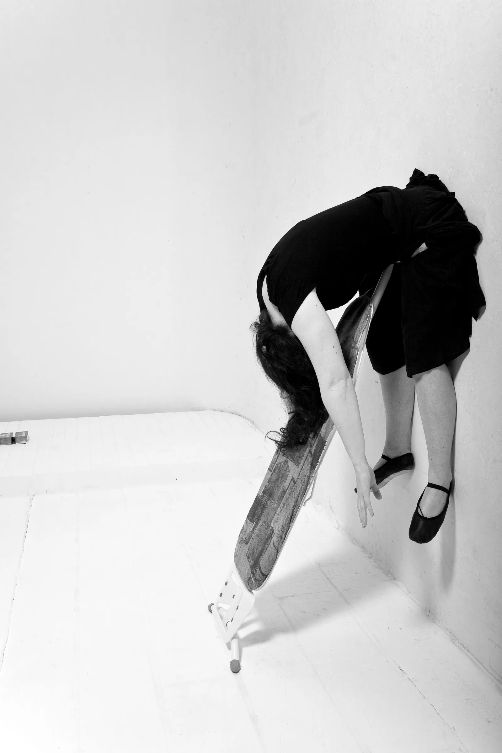 a black and white photograph by Mel Brimfield of a woman propped up and suspended against the wall by an ironing board