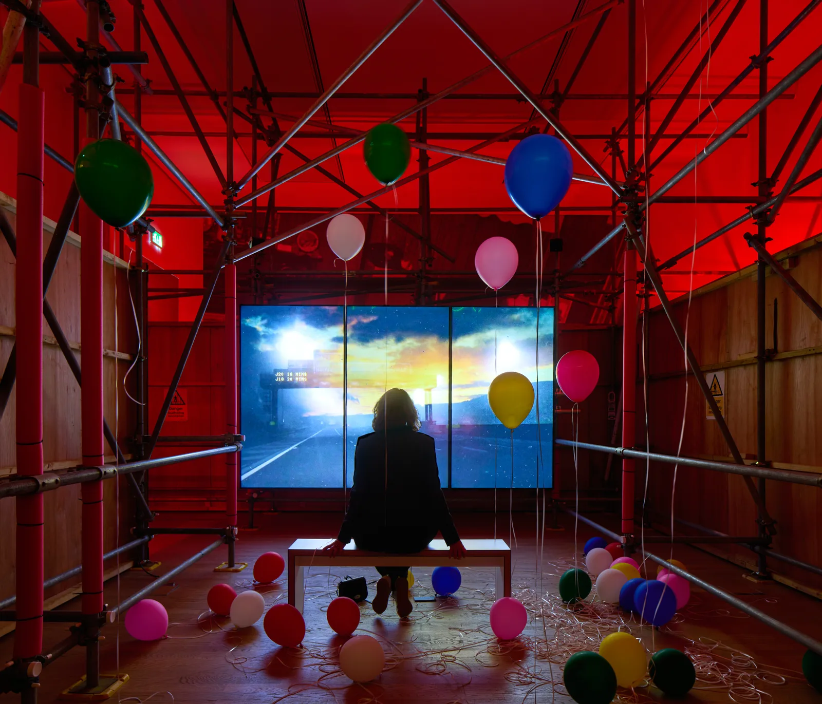 An image of a woman watching a video in a red room surrounded by balloons