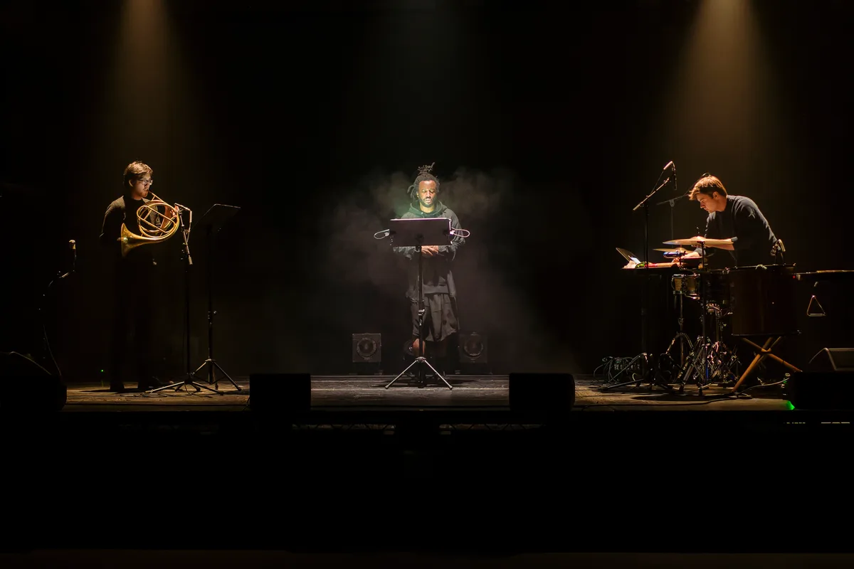 Simeon Barclay performing on a spotlit stage flanked by two musicians